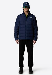 Granatowa pikowana kurtka z wysokim kołnierzem, zamek błyskawiczny i logo The North Face; w połączeniu z czarnymi spodniami oraz niebieską czapką w zielony daszek.