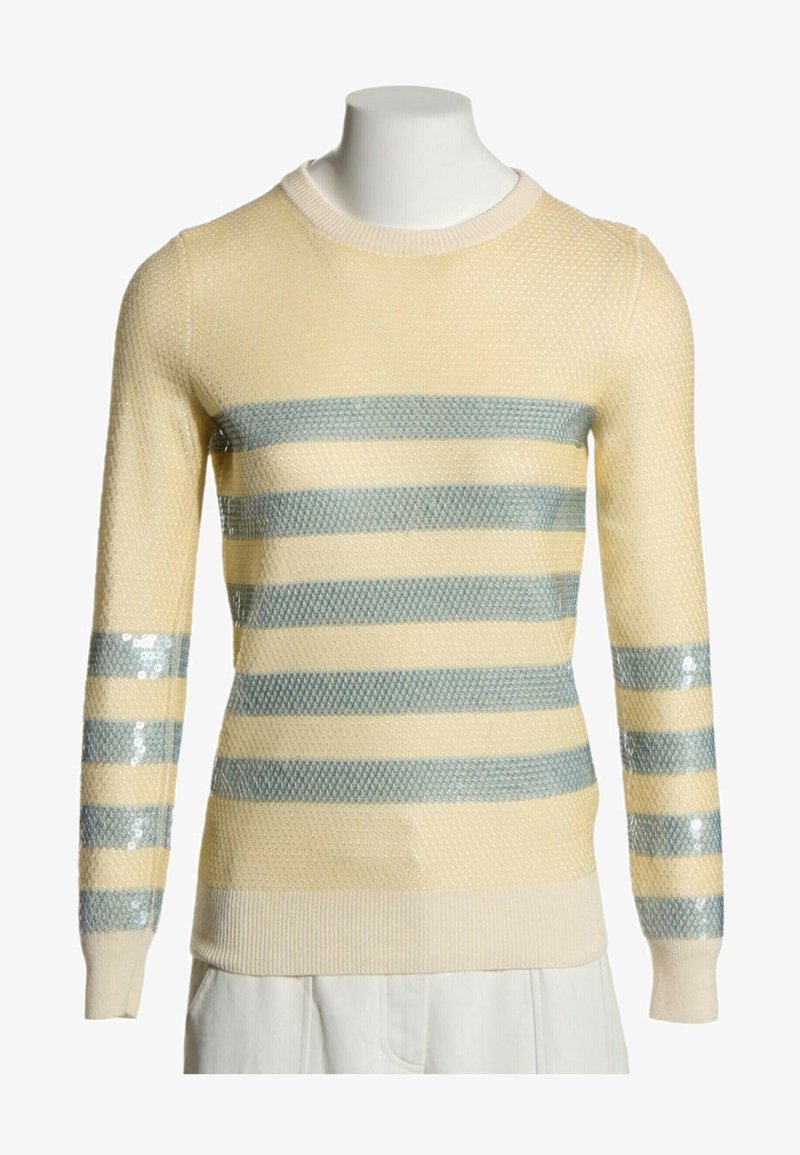 Pull en laine crème avec tissu texturé et rayures horizontales brillantes argentées-gris sur la poitrine et les manches, présenté sur un mannequin blanc.