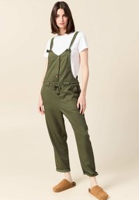 Olijfgroen overall met verstelbare bandjes, houten knopen, een trekkoord in de taille en opgerolde mouwen, gecombineerd met bruine klompen en een wit T-shirt.
