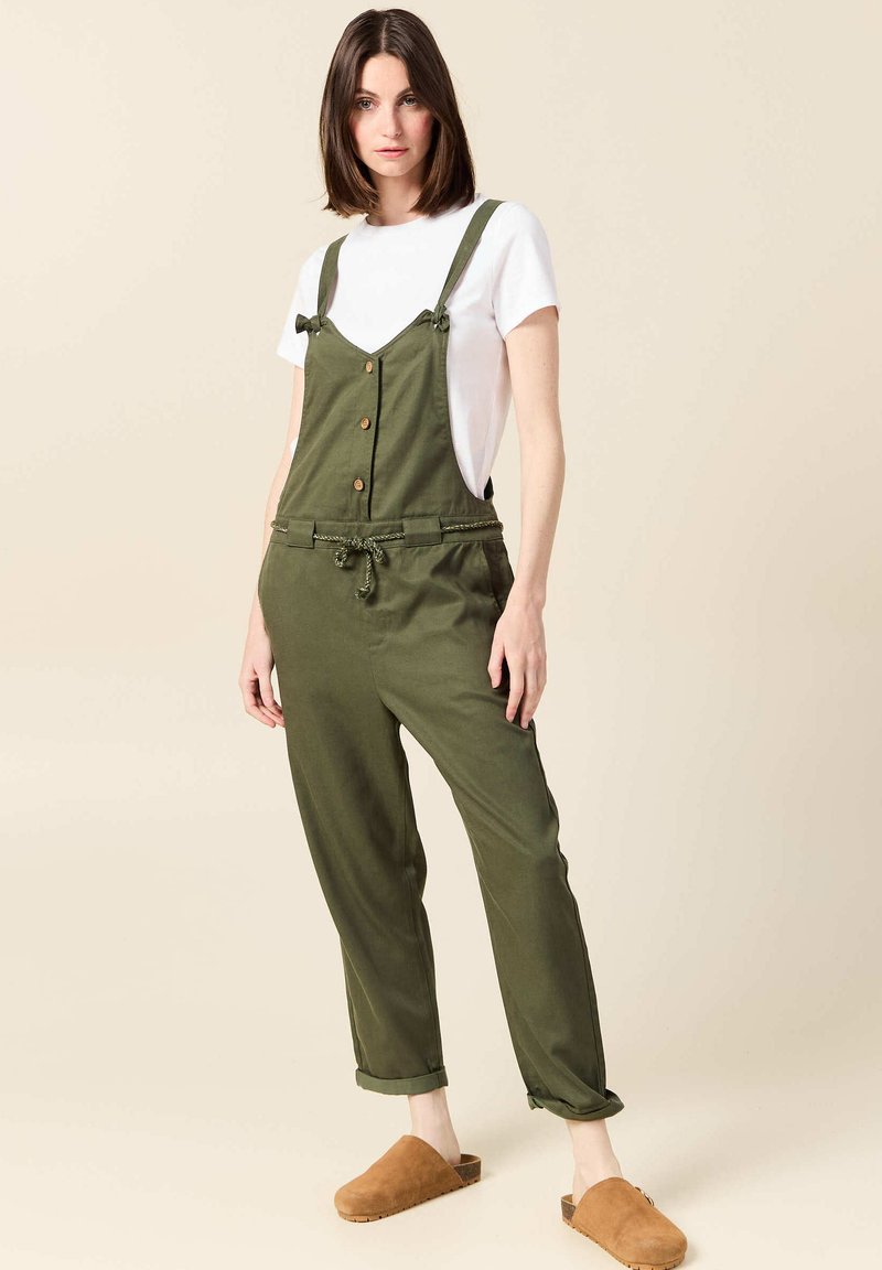 Olijfgroen overall met verstelbare bandjes, houten knopen, een trekkoord in de taille en opgerolde mouwen, gecombineerd met bruine klompen en een wit T-shirt.