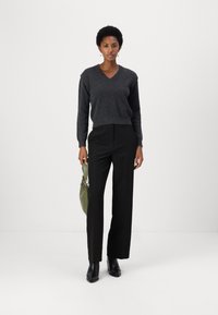 Maglione grigio a scollo a V con maniche lunghe, abbinato a pantaloni neri a vita alta. Il modello tiene una borsa verde oliva; stivali neri alla caviglia in evidenza.