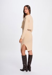 Abito lavorato a maglia beige con texture a coste, spacco laterale e maglione cropped. Abbinato a stivali lucidi marroni alti fino al ginocchio.