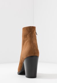 Brun ruskindsankelstøvle med høj sort hæl. Har en glat struktur, sømdetaljer og en lynlås bagpå til lukning.