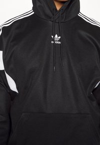 Svart Adidas-hoodie med vita tre ränder på ärmarna, vitt logo på bröstet och framficka, bärs av en person.
