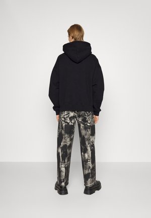 Person steht mit dem Rücken zur Kamera, trägt einen schwarzen Kapuzenpullover, schwarz-weiß gemusterte Hosen und schwarze klobige Schuhe vor einem einfarbigen Hintergrund.