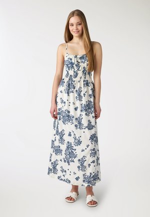 Giovane donna in piedi che indossa un abito lungo bianco senza maniche con stampe floreali blu e sandali bianchi slip-on.