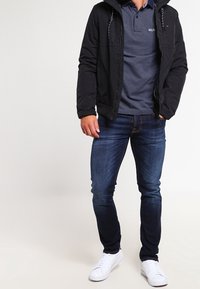 Giacca nera con cappuccio, polo blu notte con motivo testurizzato, jeans blu scuro e sneakers bianche. Dettagli includono il cordino sul cappuccio.