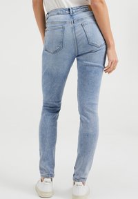 Užší modré džíny vyrobené z denimu s vysokým pasem, obsahující dvě zadní kapsy a jemné detaily šití, doplněné o bílé tenisky.