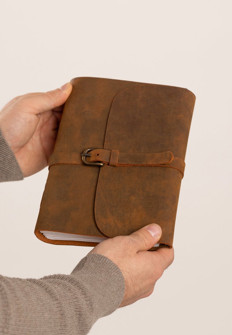 Brown leather notebook with a textured surface, secured with a buckle strap. The edges are rounded, featuring visible stitching and a crisp white interior.