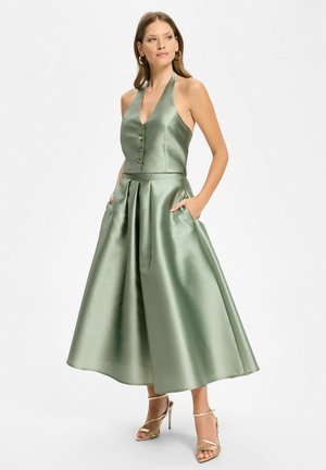 Femme portant un top dos nu en satin vert sauge avec des boutons et une jupe midi plissée assortie, mains dans les poches, talons dorés.