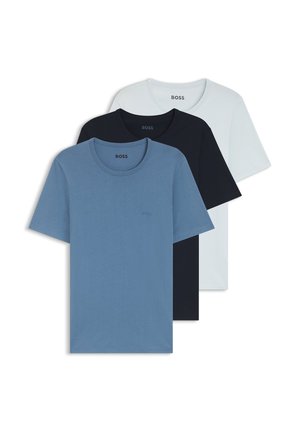 Mand stående barfodet iført en blå T-shirt med korte ærmer og sorte knælange shorts mod en ensfarvet hvid baggrund.