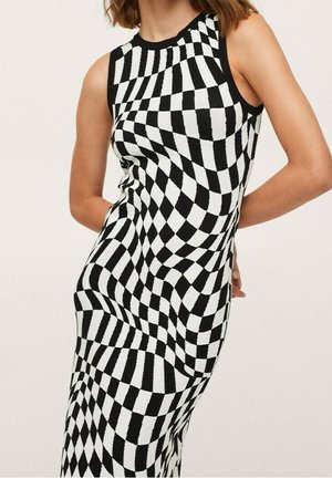 Femme portant une robe sans manches à carreaux noir et blanc avec un motif ondulé déformé, debout avec les mains dans le dos.