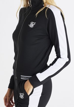 Vrouw draagt een zwart jack met rits en broek met witte strepen op de mouwen en benen, handen in de zakken, merkembleem op borst en broek.