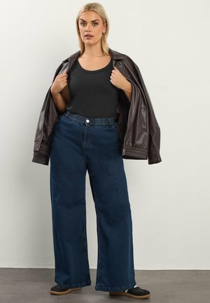 Femme blonde portant un haut noir, une veste en cuir marron foncé drapée sur les épaules, un jean large bleu et des baskets noires, debout contre un mur blanc.