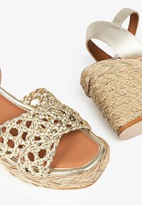 Des sandales plates en or tressées présentent une plateforme en jute tressé, des straps en cuir et une semelle intérieure texturée, alliant des éléments naturels et métalliques.