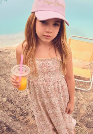 Jauna mergaitė, apsistačiusi rožinę kepuraitę ir raštuotą suknelę, laiko stiklinę apelsinų sulčių su rožiniu šiaudeliu lauke, netoli geltonos sulankstomos kėdės.
