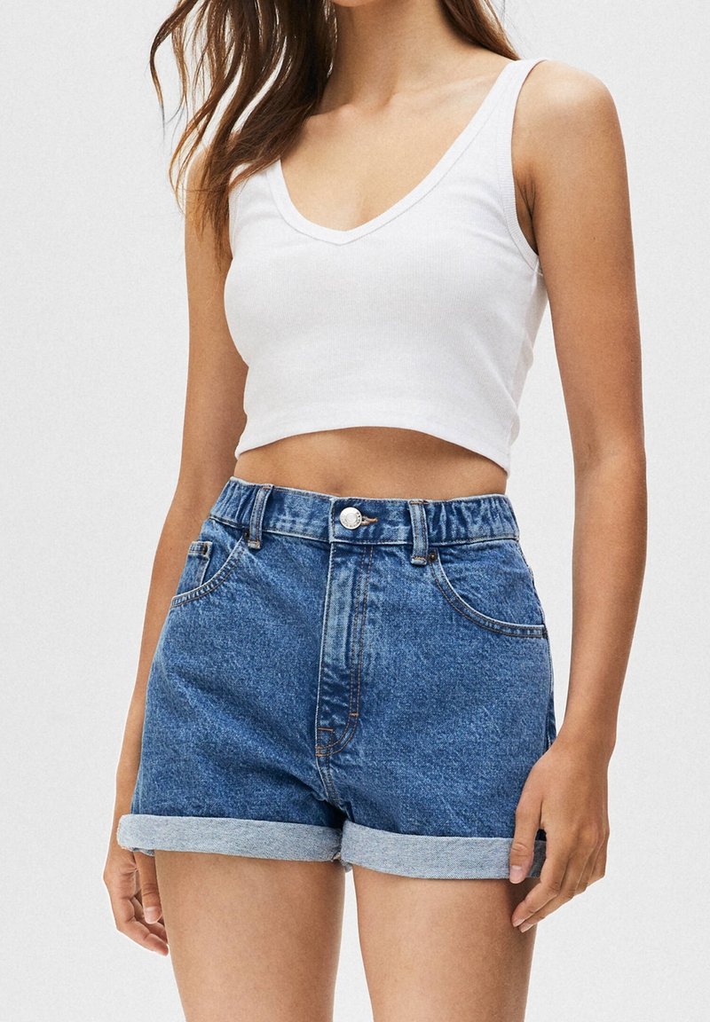 Femme portant un crop top blanc sans manches et un short en jean bleu taille haute avec revers, sur un fond uni.