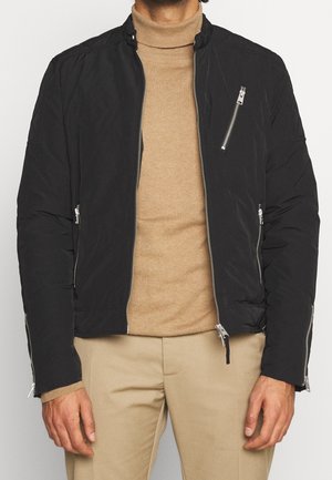 Homme portant une veste noire à fermeture éclair sur un col roulé beige et un pantalon beige, avec des poches zippées sur la poitrine et les manches.