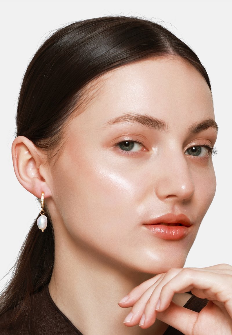 Les boucles d'oreilles en or en forme de hoop présentent une goutte en opale, mettant en valeur une surface irisée. Le mannequin a les cheveux droits et foncés, un teint lisse et une couleur de lèvres subtile.
