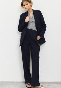 Navy tailored blazer with long sleeves, paired with high-waisted wide-leg trousers. Gray henley shirt underneath, featuring button details.