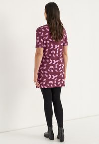 Kurzes lila Kleid mit einem pinken abstrakten Muster, kurzen Ärmeln und geradem Schnitt, kombiniert mit schwarzen Leggings und Ankle Boots.