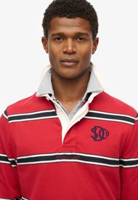 Rød og hvid stribet polo t-shirt med krave, prydet med et marineblåt logo på brystet og lavet af tekstiler i bomuldsstrik.