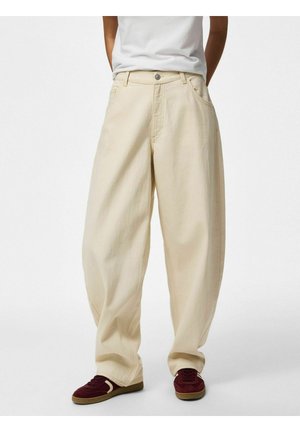 Osoba koja nosi široke bež hlače, bijelu košulju uvučenu unutra i bordo tenisice s bež potplatom, stoji ispred jednostavne pozadine.