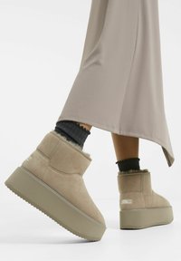 Beige kotníkové boty vyrobené z měkké velurové kůže s texturovaným svrškem a kožešinovými detaily. Mají silnou béžovou platformu pro vyšší postavu.