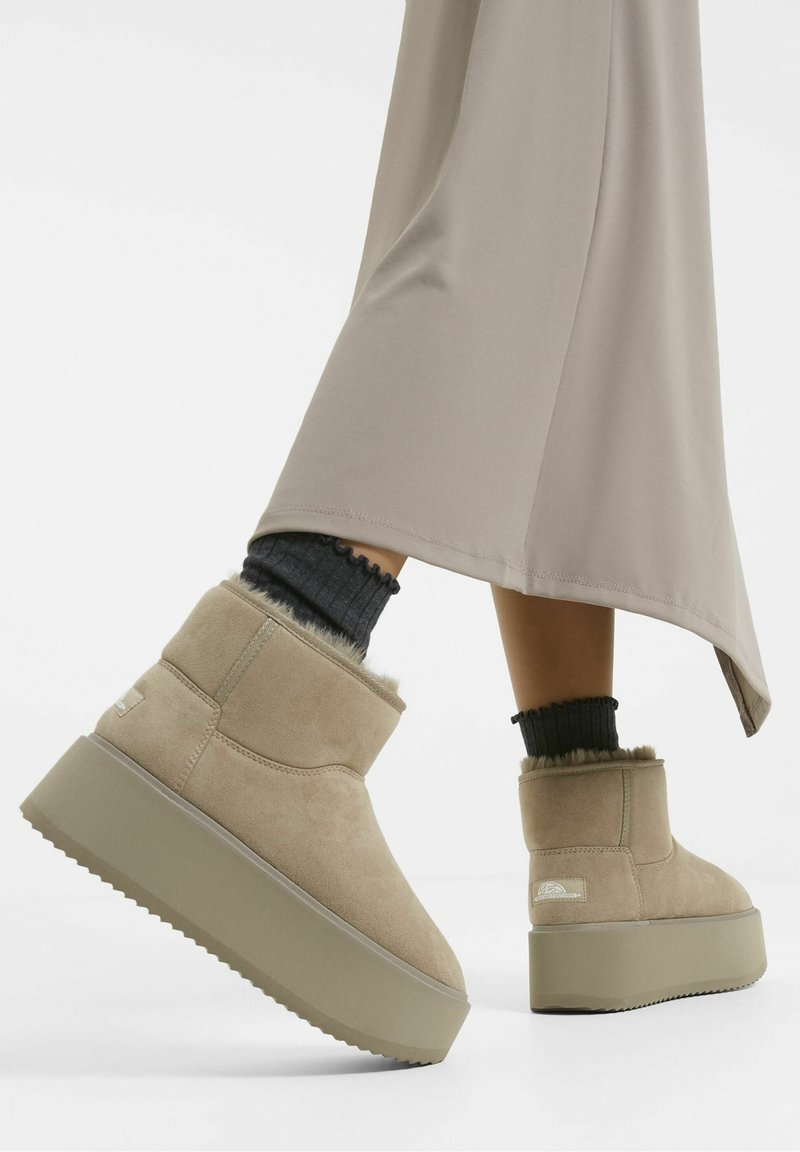 Beige kotníkové boty vyrobené z měkké velurové kůže s texturovaným svrškem a kožešinovými detaily. Mají silnou béžovou platformu pro vyšší postavu.
