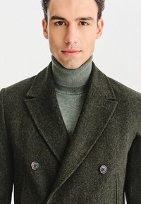 Dwurzędowy zielony płaszcz w jodełkę z wyciętym klapą, zestawiony z szarym golfem. Charakteryzuje się dużymi, okrągłymi guzikami i fakturowaną tkaniną.