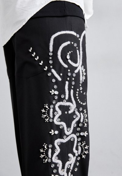 Fekete nadrág, amelyet fehér hímzett virágminták díszítenek, és gyémántszerű díszítőelemek teszik még különlegesebbé, kiemelve a textúrát és a bonyolult tervezési részleteket.