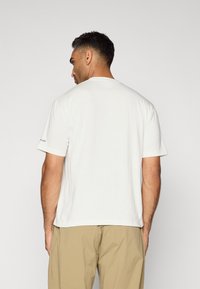 T-shirt bianco a maniche corte realizzato in cotone con scollatura rotonda. Presenta un piccolo logo decorativo sulla manica. Texture liscia.