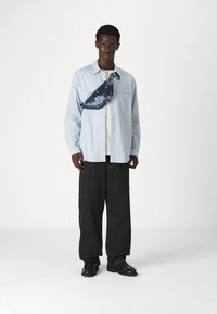 Svijetloplava košulja s dugim rukavima, crne traperice, crne cipele i fanny pack u šarenom tie-dye dizajnu nošen preko grudnog koša. Jednostavan, opušten kroj.