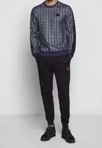 Sudadera azul marino con patrón repetido del logo de EA7, combinada con pantalones negros que presentan el logo dorado de EA7, y zapatillas negras.