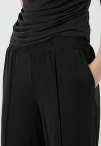 Close-up van een persoon die losse zwarte broek draagt met een elastische tailleband en zijzak, met een hand gedeeltelijk in de zak.
