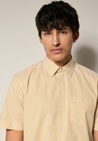 Camisa de manga corta en amarillo claro con un patrón geométrico de diamantes. Cuenta con un collar y cierre de botones, textura de tela suave.