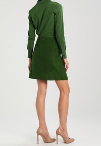 Blusa verde entallada con manga larga combinada con una falda de ante verde. La modelo lleva tacones nude. Tejido liso, diseño ajustado.