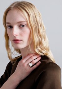 Hõbedane südamekujuline sõrmus käel, mida kantakse pruuni tekstuuriga ülikonna peal. Juuksed on blondid ja lainelised, heleda halli tausta ees.