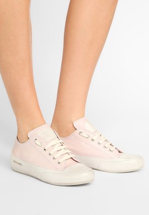 Personne portant des baskets basses rose clair avec des semelles et des lacets blancs, debout sur une surface unie gris clair.