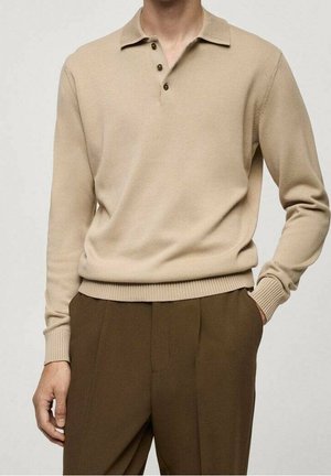 Polo de punto beige de manga larga con tres botones, combinado con pantalones marrones de talle alto, una mano en el bolsillo, fondo neutro.