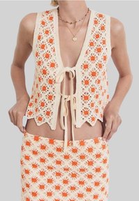 Háčkovaný krémový komplet vesty a sukně s oranžovými květinovými vzory. Vesta má zavazování na uzel a vlnité lemy; oba kusy mají texturovaný, otevřený vzor.