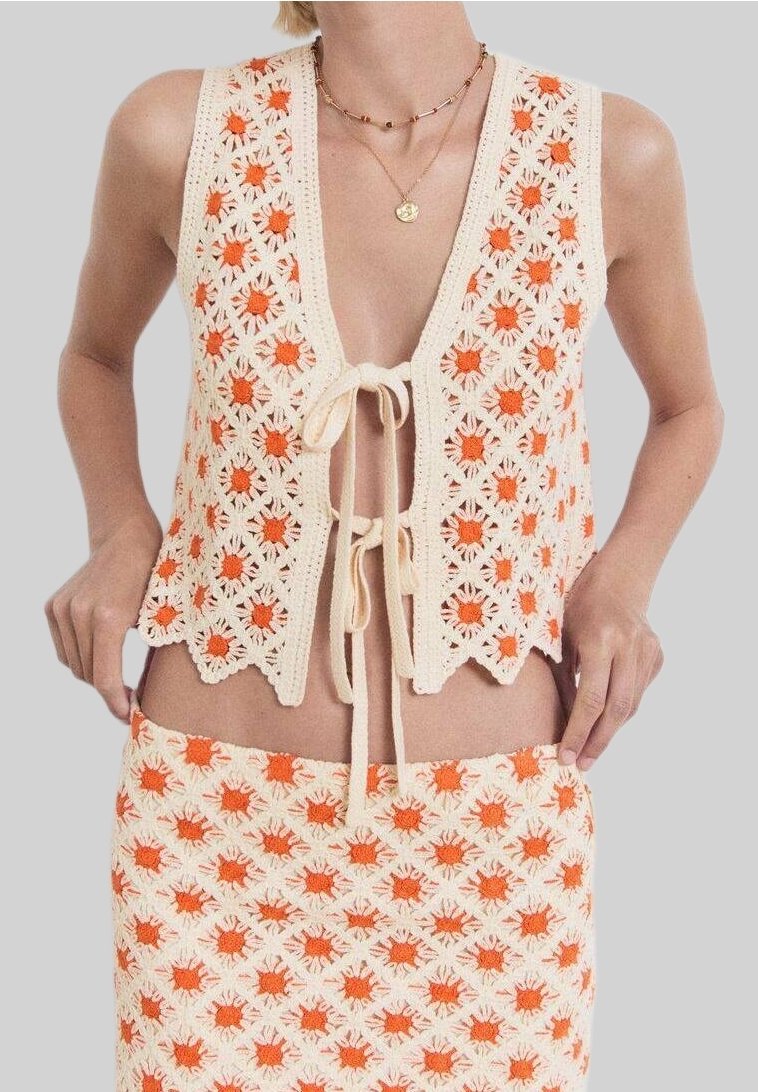 Háčkovaný krémový komplet vesty a sukně s oranžovými květinovými vzory. Vesta má zavazování na uzel a vlnité lemy; oba kusy mají texturovaný, otevřený vzor.