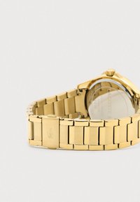 Gold metal link watch band with clasp featuring engraved crocodile logo, and partially visible round watch face angled away.