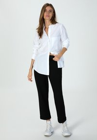 Weißes Hemd mit langen Ärmeln und einer Brusttasche, kombiniert mit schwarzen tapered Hosen und weißen Sneakers, auf einem neutralen Hintergrund präsentiert.