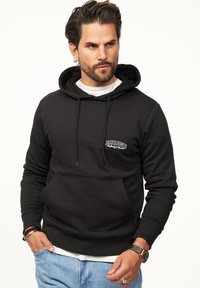 Schwarzer Pullover-Hoodie mit einer Kängurutasche, Kordelzugkapuze und weißem Logo auf der Brust. Über einem weißen Shirt getragen und mit blauen Jeans kombiniert.