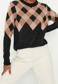 Personne portant un pull noir avec un motif losange rose et marron sur la poitrine et le haut des bras, associé à un pantalon blanc cassé.