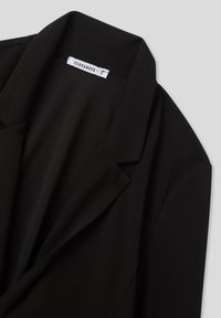 Blazer nero su misura con colletto a punta e etichetta bianca che mostra il marchio "Terranova" e la taglia "S" su uno sfondo grigio chiaro.