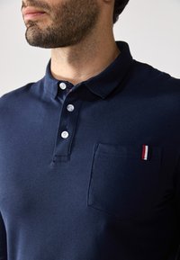 Polo de color marino hecho de un material texturizado; cuenta con un cuello, tres botones blancos y un bolsillo en el pecho con un acento rojo, blanco y azul.