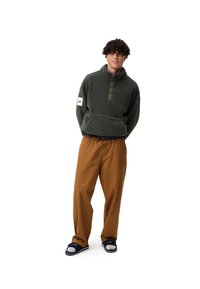Pull en polaire vert foncé avec un col zippé et trois boutons, associé à un pantalon ample marron et des sandales à glissière bleu marine.