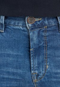 Jeans in denim blu con una texture strappata, chiusura a bottone di rame e cuciture gialle sulla vita e sulle tasche.